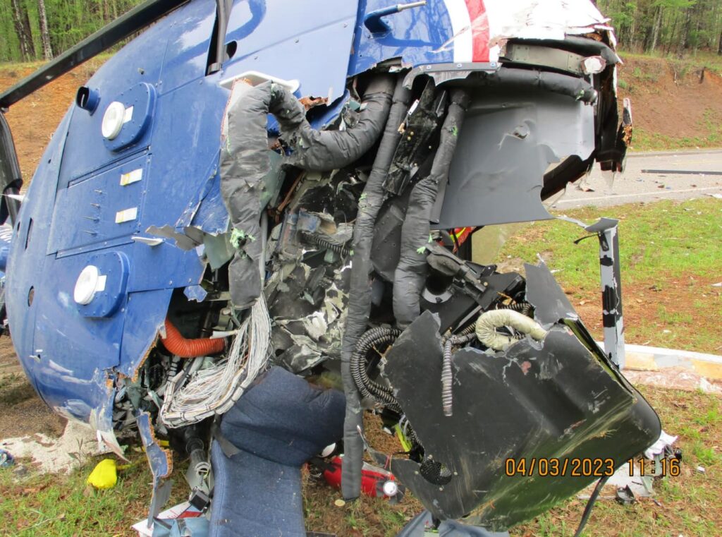 Close-up of the damaged lower fuselage and internal wiring of the helicopter, showing torn panels, exposed cables, and shattered avionics components amid crash debris.